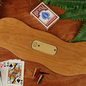 May include: A smooth, light brown wooden cribbage board with an organic, wavy shape. The board features a central rectangular brass inlay with two small holes. A deck of Bicycle playing cards, four playing cards, and five small pegs are included.