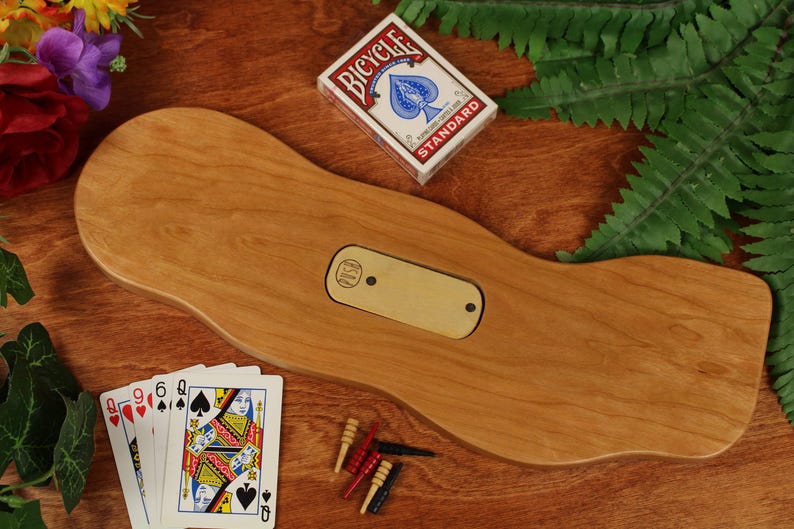May include: A smooth, light brown wooden cribbage board with an organic, wavy shape. The board features a central rectangular brass inlay with two small holes. A deck of Bicycle playing cards, four playing cards, and five small pegs are included.