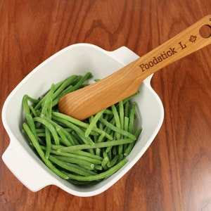 Peut inclure: Un bol en céramique blanche rempli de haricots verts et une spatule en bois avec le texte "Foodstick L" et un logo de feuille d'érable.
