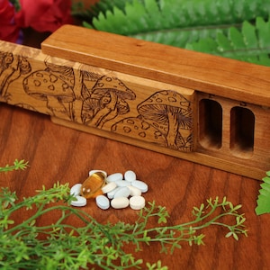 May include: A wooden box with a sliding lid, featuring a laser-etched design of mushrooms. The box has three compartments for storing pills or other small items. The box is made of light brown wood and has a natural finish.