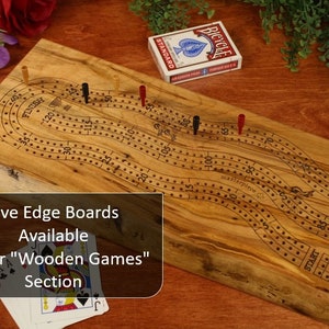 May include: A rectangular live edge wooden cribbage board featuring a winding track with numbered holes from start to finish. The board includes red, black, and yellow pegs, a deck of Bicycle playing cards, and text reading Live Edge Boards Available In Our Wooden Games Section.