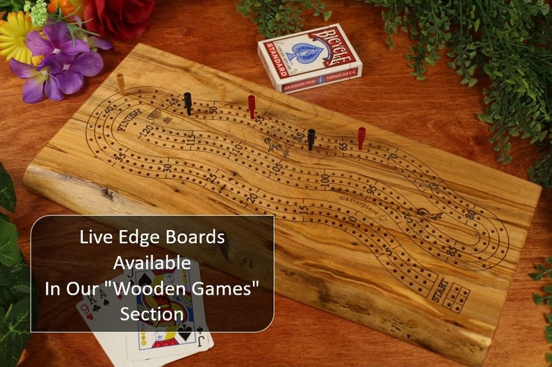 May include: A rectangular live edge wooden cribbage board featuring a winding track with numbered holes from start to finish. The board includes red, black, and yellow pegs, a deck of Bicycle playing cards, and text reading Live Edge Boards Available In Our Wooden Games Section.