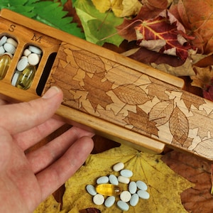 May include: A wooden pill box with a sliding lid, featuring a leaf design. The box has two compartments labelled "S" and "M". The box is open and there are pills inside.