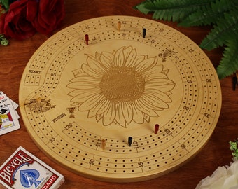 Könnte beinhalten: Ein Cribbage-Brett aus Holz mit einem Sonnenblumen-Design. Das Brett ist rund und aus hellem Holz gefertigt. Die Sonnenblume befindet sich in der Mitte des Bretts und ist von einer Reihe von Löchern für Stifte umgeben. Das Brett ist am Rand nummeriert, beginnend bei 0 und bis 120.
