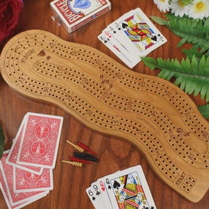May include: A light brown wooden cribbage board with a wavy, elongated shape featuring numbered tracks from start to finish. The board includes small red, black, and yellow metal pegs, along with several playing cards scattered around the surface.