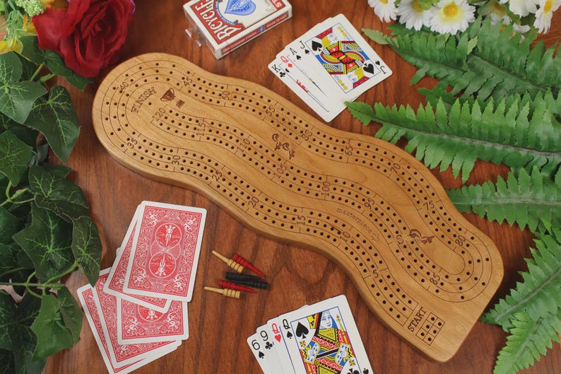 May include: A light brown wooden cribbage board with a wavy, elongated shape featuring numbered tracks from start to finish. The board includes small red, black, and yellow metal pegs, along with several playing cards scattered around the surface.