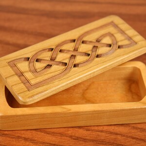 May include: A wooden box with a hinged lid. The lid features a carved Celtic knot design. The box is made of light wood and has a natural finish.