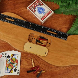 May include: A light brown wooden cribbage board with a central storage compartment containing small pegs. A black 12-inch ruler is placed across the board. A deck of Bicycle playing cards and several loose red, black, and yellow pegs are visible.