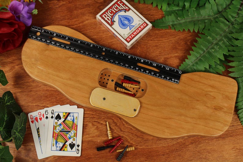 May include: A light brown wooden cribbage board with a central storage compartment containing small pegs. A black 12-inch ruler is placed across the board. A deck of Bicycle playing cards and several loose red, black, and yellow pegs are visible.