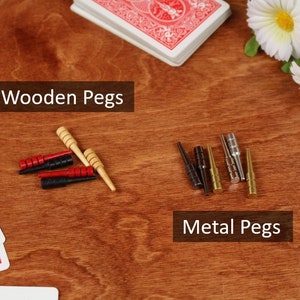 May include: A collection of game pegs including wooden pegs in red, black, and natural tan, and metal pegs in black, gold, and silver finishes. The image features text labels reading Wooden Pegs and Metal Pegs alongside playing cards.
