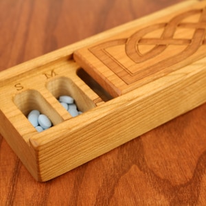 May include: Wooden pill box with a Celtic knot design on the lid. The box has three compartments labeled "S", "M", and "L" and is filled with blue pills.