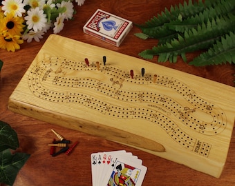 May include: A wooden cribbage board with a natural wood finish. The board has a curved track with numbered spaces and a "Start" space. There are several pegs on the board and a deck of playing cards nearby.