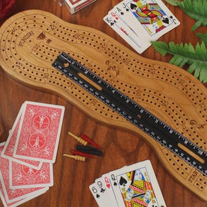 May include: A light brown wooden cribbage board with a curved shape, featuring numbered tracks and a finish line. A black ruler placed diagonally across the board shows a length of approximately 12 inches. Red, black, and yellow pegs and playing cards are included.