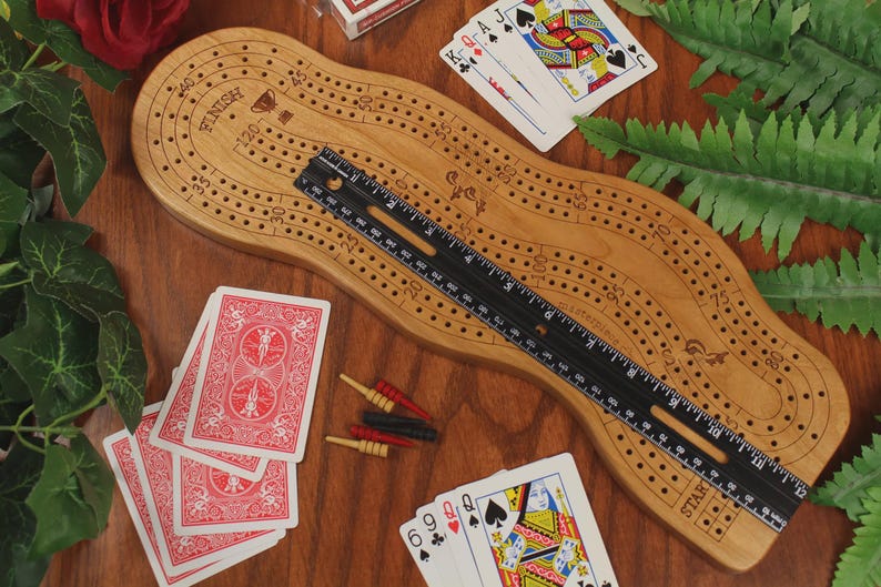 May include: A light brown wooden cribbage board with a curved shape, featuring numbered tracks and a finish line. A black ruler placed diagonally across the board shows a length of approximately 12 inches. Red, black, and yellow pegs and playing cards are included.