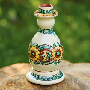 Puede incluir: Una botella decorativa de cerámica con un diseño floral. La botella es de color crema con detalles en amarillo, marrón y turquesa. Mide aproximadamente 15 cm de altura y tiene una base acampanada y una parte superior redonda.