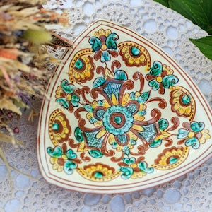 Könnte beinhalten: Eine dreieckige Keramikschale mit floralem Design. Die Schale hat eine cremefarbene Basis mit einem braunen Rand und ist mit türkisfarbenen, gelben und braunen Blumenmustern verziert. Die Schale wird wahrscheinlich zum Servieren oder Ausstellen verwendet.
