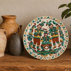 Op de afbeelding: Een decoratieve keramische schaal met een geschulpte rand, met een kleurrijk volkskunstontwerp van drie figuren in traditionele kledij. De schaal is omgeven door vazen en een plant.