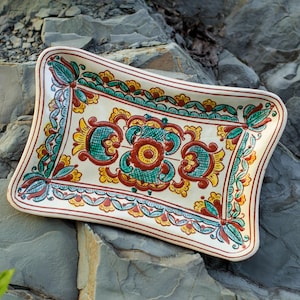 Puede incluir: Una bandeja rectangular de cerámica con base color crema que presenta un patrón simétrico pintado a mano en tonos verde azulado, rojo óxido y amarillo. El diseño incluye motivos florales y volutas intrincadas en el borde y centro.
