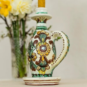 Puede incluir: Un candelabro de cerámica con asa, con un diseño floral en amarillo, verde y marrón. El candelabro tiene una base blanca y una parte superior decorativa. Una vela amarilla está insertada en la parte superior. El soporte mide unos 15 cm de alto.