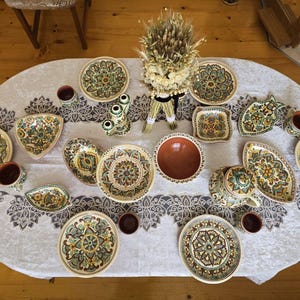 Op de afbeelding: Een verzameling handbeschilderd keramiek serviesgoed, waaronder borden, kommen en kopjes, gerangschikt op een wit tafelkleed. De stukken hebben een bloemmotief in groen, geel en bruin. Een decoratieve tarwebundel staat in het midden.