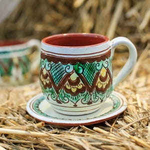 Könnte beinhalten: Ein Keramikbecher- und Untertassen-Set mit traditionellem Volkskunst-Design. Der Becher hat eine weiße Basis mit braunen, grünen und gelben dekorativen Mustern. Der Innenraum ist rot. Das Set steht auf Stroh.
