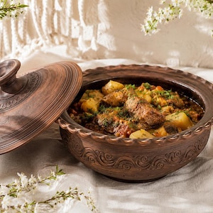 May include: A dark brown clay pot with a matching lid, featuring intricate embossed swirl patterns along the rim. The pot is filled with a hearty stew containing chunks of meat, potatoes, and vegetables, garnished with fresh green herbs.