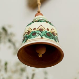 Op de afbeelding: Een keramische bel met een bruin interieur en een crèmekleurige buitenkant, versierd met groene en bruine bloempatronen. De bel hangt aan een natuurlijk gekleurd touw en toont een rustieke esthetiek.