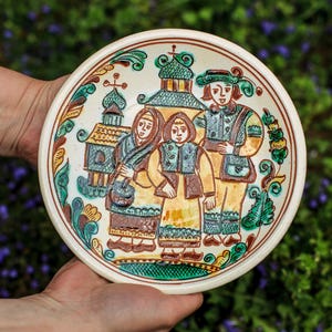 May include: A hand-painted ceramic plate with a cream-colored base and a detailed folk art design. The artwork depicts three figures in traditional clothing, a building, and decorative floral elements in green, brown, and yellow.