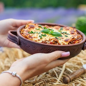 May include: A baked lasagna dish in a brown ceramic bowl with handles, garnished with fresh basil. The lasagna is held by hands, with a blurred background of greenery and straw.