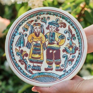 Op de afbeelding: Een ronde, handbeschilderde keramische schotel met een volkskunstontwerp. De schotel toont twee figuren in traditionele kledij, omringd door bloemen- en krulmotieven in geel, blauw en bruin op een witte achtergrond.