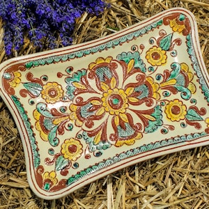 Könnte beinhalten: Rechteckige Keramikservierplatte mit cremefarbenem Grund, verziert mit einem Blumenmuster in Gelb, Braun und Türkis. Die Platte hat einen gewellten Rand und liegt auf einem Hintergrund aus Stroh und Lavendel.