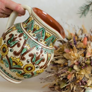 Op de afbeelding: Een handbeschilderde keramische kan met een crèmekleurige basis en ingewikkelde bloempatronen in bruin, geel en groen. De kan heeft een bruin interieur en een gebogen handvat. Het ontwerp doet denken aan volkskunst.