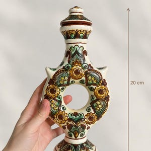 Può includere: Un recipiente in ceramica color crema con un'apertura circolare centrale, decorato con motivi floreali dipinti a mano in marrone, verde e giallo. La bottiglia misura 11 cm di larghezza e 20 cm di altezza, con tappo e base decorativi.