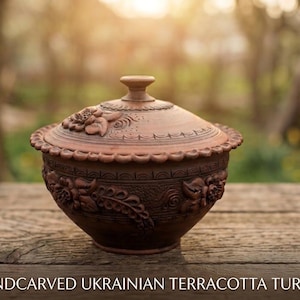 May include: A hand-carved terracotta tureen with a lid. The tureen is brown with floral and leaf designs. The lid has a knob and is decorated with carved flowers. The tureen sits on a wooden surface.