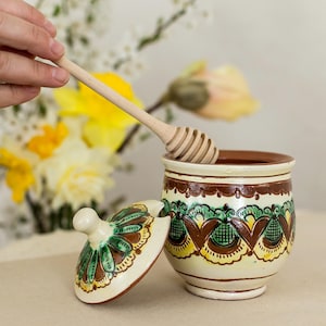 May include: A ceramic honey pot with a decorative lid and a wooden honey dipper. The pot is cream-colored with brown and green floral designs. The lid is off to the side, and the dipper is in the pot.