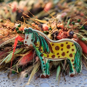 May include: A ceramic horse figurine with a yellow body and brown spots, green and brown mane, and a white face. The horse is set against a backdrop of dried flowers and grasses.