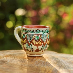 Pode incluir: Uma caneca de cerâmica pintada à mão com base creme e padrões intrincados em verde, castanho e amarelo. O interior é vermelho e a caneca tem uma pega curva. A caneca está colocada numa superfície de madeira.