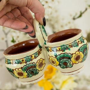 Puede incluir: Un recipiente de cerámica con doble asa y dos compartimentos redondos. La cerámica de color crema está decorada con motivos florales amarillos y verdes. El interior de las tazas es marrón. El recipiente lo sostiene una persona.
