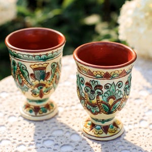 May include: Two ceramic goblets with a cream base and a red interior. The goblets feature intricate floral and bird designs in green, yellow, and brown. The goblets are placed on a white lace tablecloth.