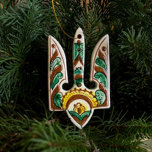 Op de afbeelding: Een keramische kerstversiering in de vorm van een drietand, met een traditioneel Oekraïens ontwerp. De versiering is beschilderd met bruine, groene en gele bloempatronen op een witte achtergrond. Het hangt tegen een achtergrond van groene dennennaalden.