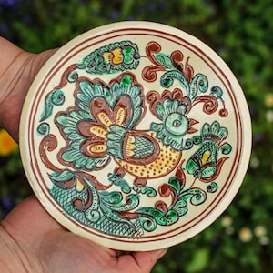 Puede incluir: Un plato redondo de cerámica pintado a mano con un diseño de pájaro y flores. El plato tiene una base de color crema con un borde marrón y está decorado con detalles verdes, marrones y amarillos. El plato está sostenido en dos manos.