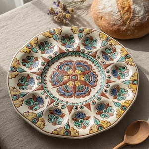 Può includere: Un piatto decorativo in ceramica con base color crema e un colorato motivo floreale. Il piatto presenta un motivo centrale a stella e scomparti individuali. Sono visibili anche una pagnotta e un cucchiaio di legno.