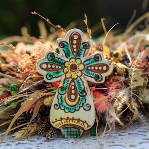May include: A ceramic cross with a floral design in yellow, teal, and brown, set against a backdrop of dried flowers and foliage. The cross is hand-painted with intricate details and stands out with its vibrant colors.