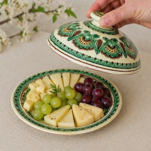 May include: A decorative ceramic serving dish with a lid, featuring a green and brown floral design. The dish holds grapes and cheese. The lid is being lifted to reveal the contents. The dish is approximately 8 inches in diameter.