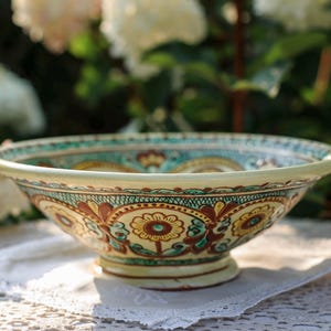 Op de afbeelding: Een decoratieve keramische kom met een brede rand en een voetstuk. De kom heeft een handgeschilderd bloemmotief in tinten geel, bruin en blauwgroen tegen een crèmekleurige achtergrond. De kom staat op een wit kanten tafelkleed.