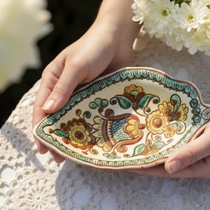 May include: A hand-painted, ceramic leaf-shaped dish. The dish features a cream-colored base with floral designs in yellow, brown, and teal. The rim is outlined in teal. The dish is held by two hands, with a bouquet of white flowers in the background.
