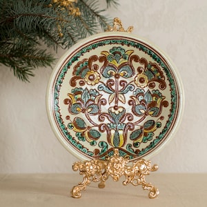 Puede incluir: Un plato decorativo de cerámica con un diseño floral en marrón, azul y amarillo, exhibido sobre un soporte de metal dorado. El plato tiene un fondo color crema y un borde verde. Un jarrón a juego es visible a la izquierda.
