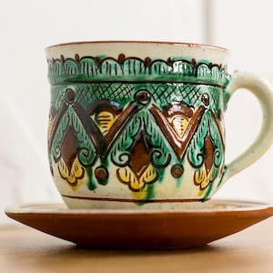 May include: A ceramic mug and saucer set with a hand-painted design. The mug features a green, brown, and yellow geometric pattern. The handle and rim are off-white. The saucer is a solid brown color. The set is likely handmade.
