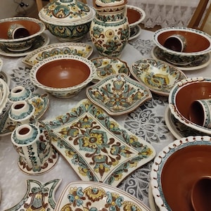 Puede incluir: Una colección de vajilla de cerámica con un diseño floral en tonos marrones, amarillos, azules y verdes. El juego incluye cuencos, platos, tazas y fuentes, todos con un patrón decorativo similar. Las piezas están dispuestas sobre un mantel blanco.