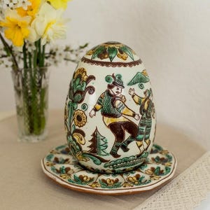 Puede incluir: Un adorno de cerámica con forma de huevo sobre un plato circular a juego, con ilustraciones de arte popular pintadas a mano de figuras con vestimenta tradicional, pinos y motivos florales en tonos verde, marrón, amarillo y crema.